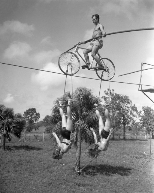Man riding a bicycle on a tightrope while two performers hang from its tires