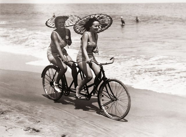 Two women in swimwear enjoy a tandem bike ride down the beach with parasols in hand. (Photo Credit: George Rinhart/Corbis via Getty Images)