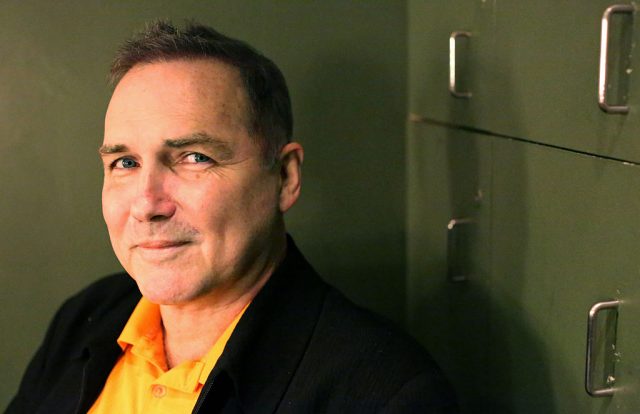 Norm Macdonald sitting beside a cabinet