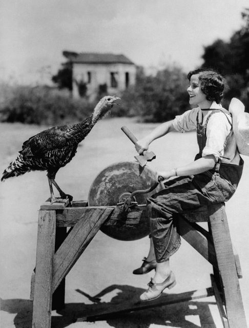 Barbara Kent sharpening an axe while a turkey stands in front of her
