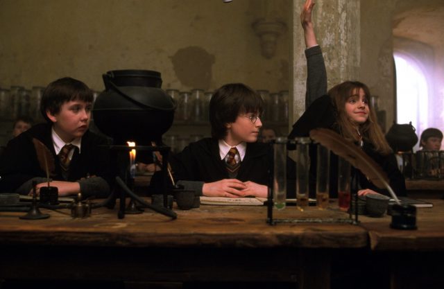 Neville Longbottom and Harry Potter looking at Hermione Granger as she raises her hand during their potions lesson