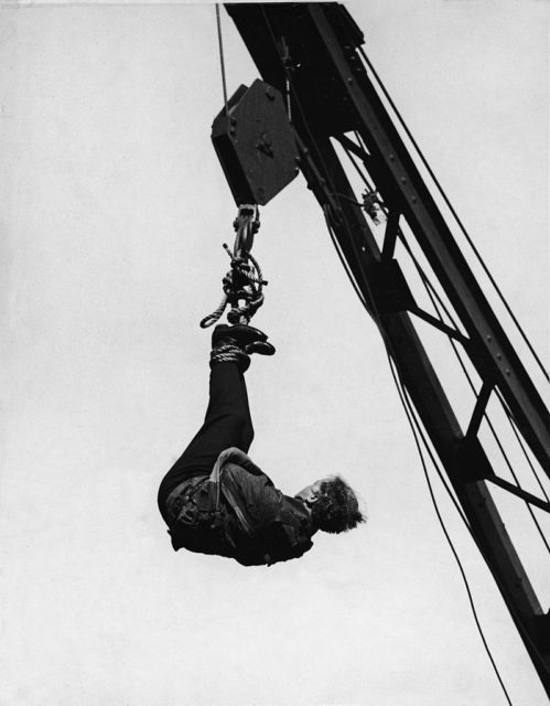 Houdini wears a straightjacket and hangs from a crane during an escape act