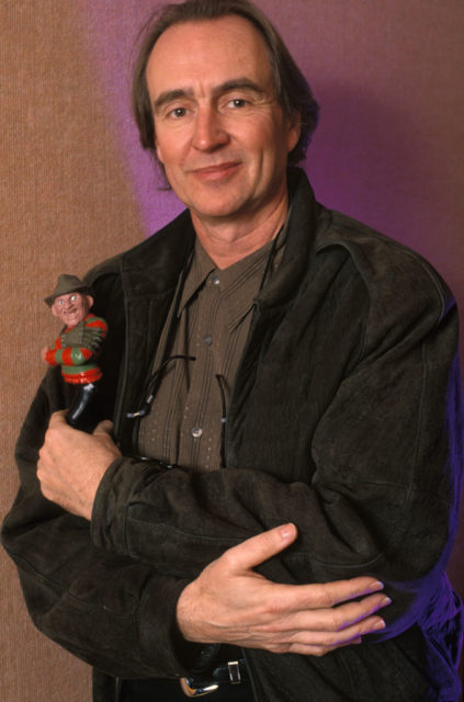 Wes Craven leans against a wall and holds a Freddy Krueger figurine