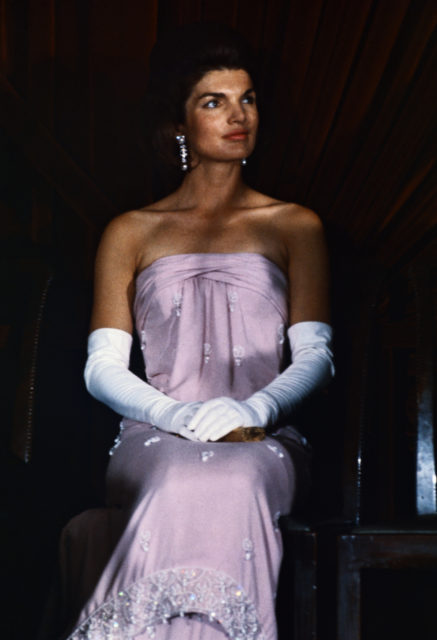 Jackie Kennedy sits in pink dress with white gloves and dangle earrings.