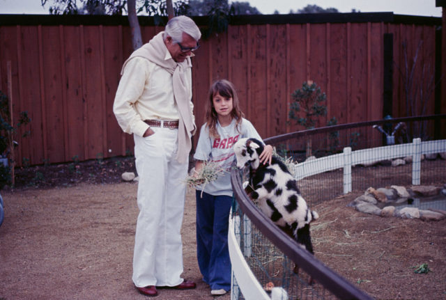 Cary Grant stands with young daughter Jennifer Grant while she pets a goat.
