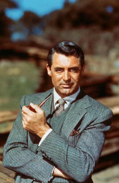 Young Cary Grant poses in a suit holding a smoking pipe. 
