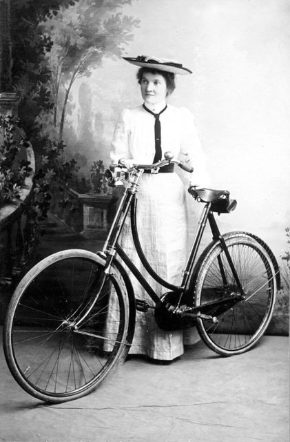 A young woman in a long dress posing behind a bicycle in 1900