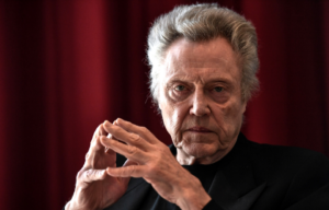 Christopher Walken sitting in front of red curtain with his fingers touching, looking at the camera