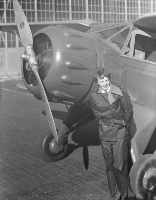Amelia Earhart beside the propeller of her plane in pilot gear.