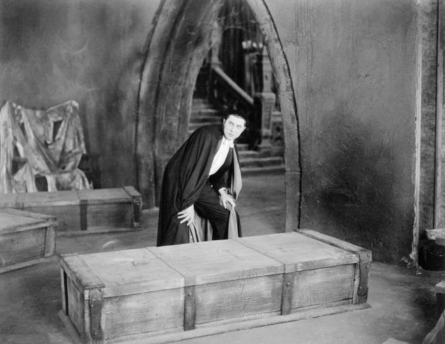 Bela Lugosi dressed as Dracula leans over a coffin. 