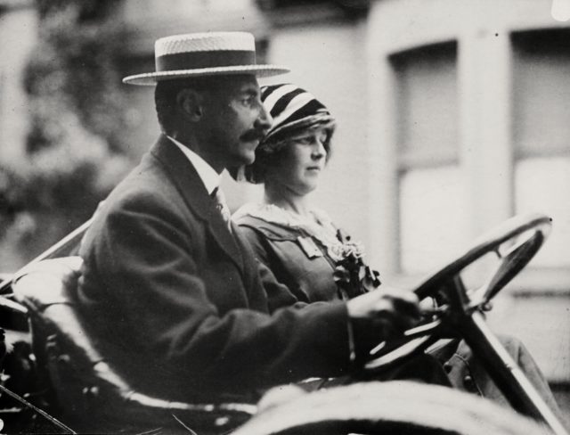 John Jacob Astor drives an automobile and Madeleine Force sits in the passenger seat.