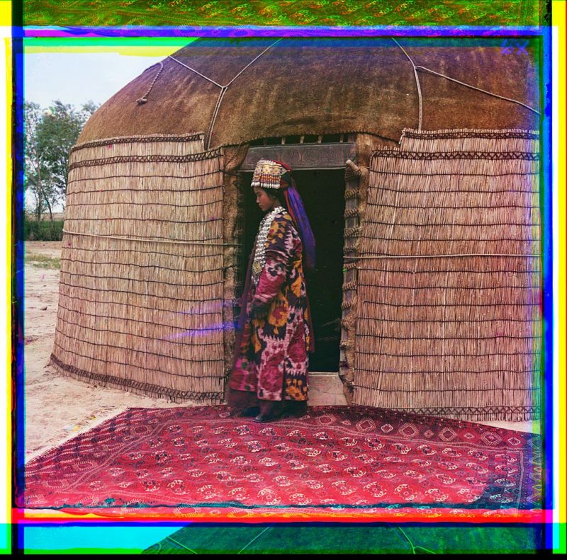 [Full-length profile portrait of a woman, possibly Turkman or Kirgiz, standing on a carpet at the entrance to a yurt, dressed in traditional clothing and jewelry]