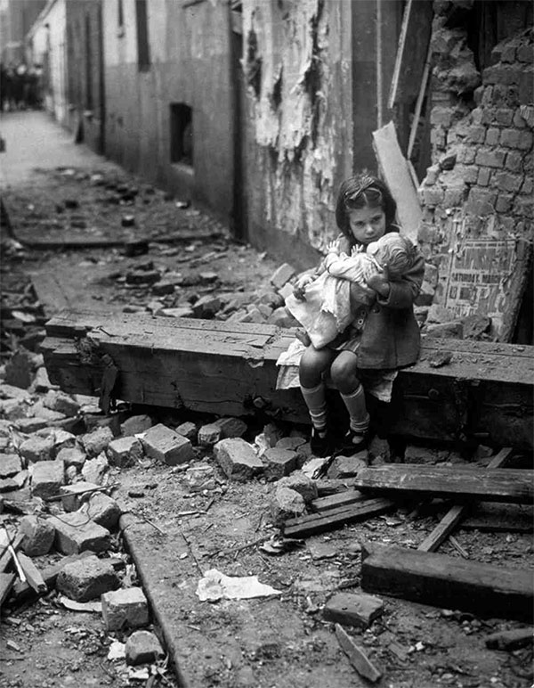 girl holds her doll while standing among the rubble of her home which was damaged by bombs in 1940