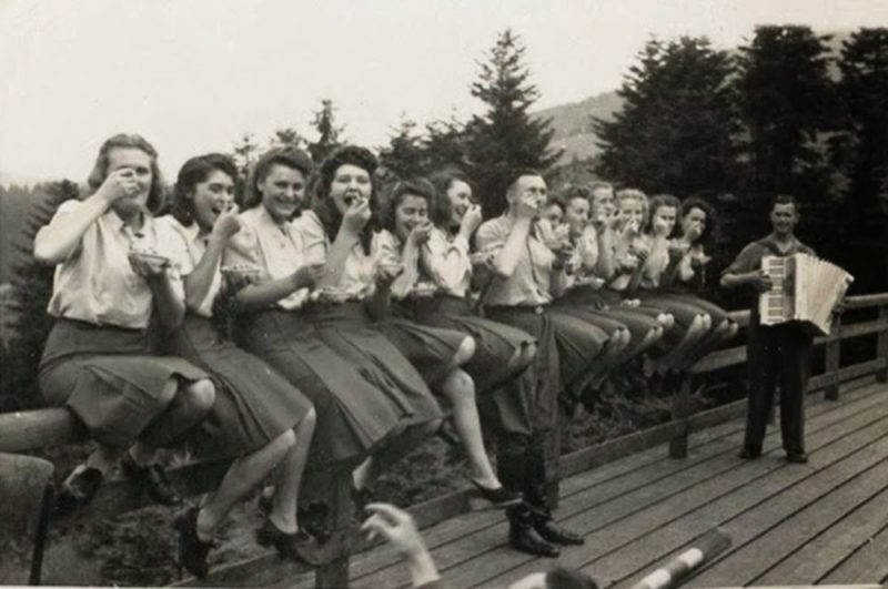Twelve SS auxiliaries sit happily on a fence railing eating blueberries given to them by an SS officer.