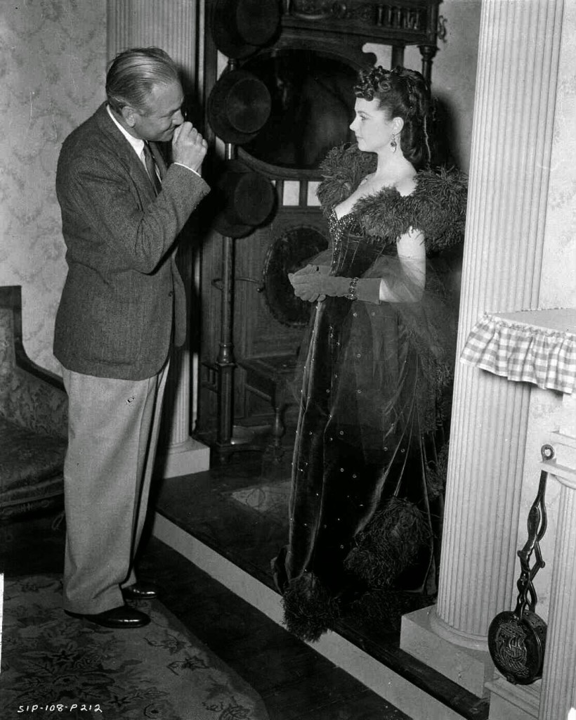 Victor Fleming looking at Vivien Leigh as Scarlett O'Hara, who is standing in doorway, wearing red gown.