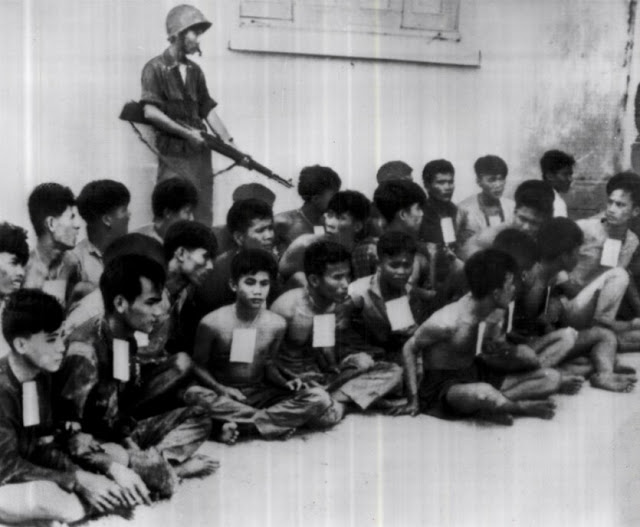 October 5, 1964 featuring partially stripped and tied Viet Cong prisoners sit on ground under armed guard in prison camp