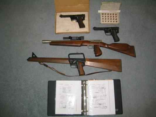 The Gyrojet Family, which are two Gyrojet pistols, a carbine, and the rifle. In the top-right corner is a box of 13 mm rockets, and at the bottom is a diagram book for the guns. source