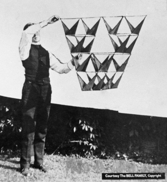 A view of one of the project workman holding an early design of the tetrahedron kite, with opposing triangular vanes, photographed outside against a white sheet.