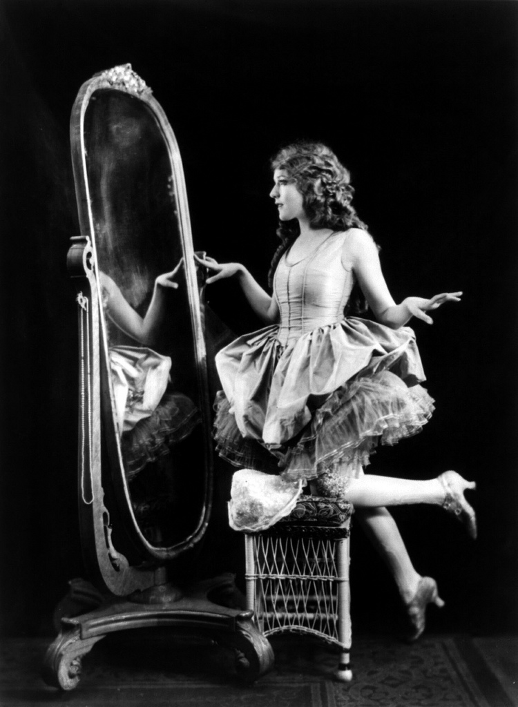 Mary Pickford, full length, facing left; kneeling on chair in front of mirror. .Source
