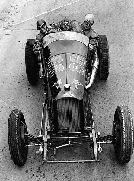 Posed behind the wheel of his racer is driver Ralph De Palma and to his left is his mechanic, Lindley Bothwell. (Photo Credit: Lew Nichols Photography / Wikimedia Commons / Public Domain)
