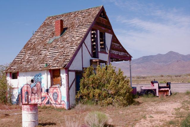 Rear view of the Santa Claus Land Sales Office looking northeast. To the right is the Old 1225, a derailed, pink children's train tagged with graffiti. U.S. Route 93, Ithaca Peak (left), and Turquoise Mountain (right) can be seen in the background. Source