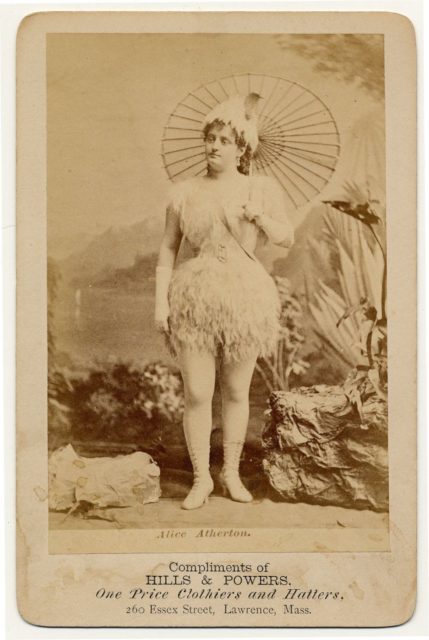 Alice Atherton in a short, feathery costume, tights, parasol, mid-forearm white gloves, mid-calf white heeled and laced boots, feathery hat.
