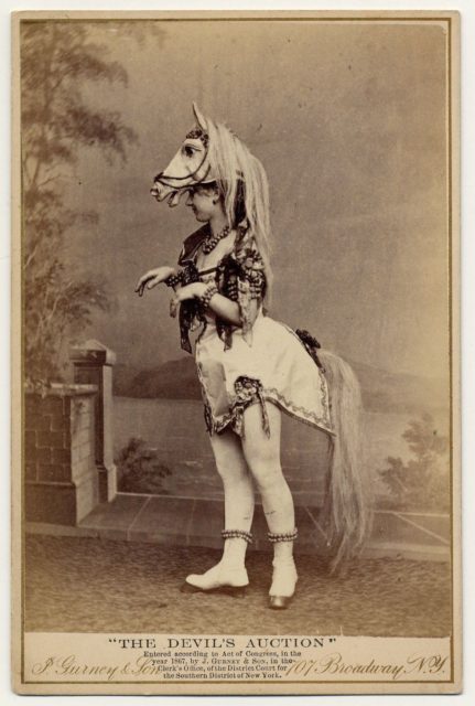 Eliza Blasina wearing horse-head headdress, short costume with attached horsetail, rows of round beads or bells around ankles, wrists, neck and upper arm.