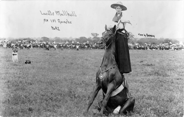 Lucille Mulhall, a Wild West performer here captured on camera as she stands on a horse that is seated.