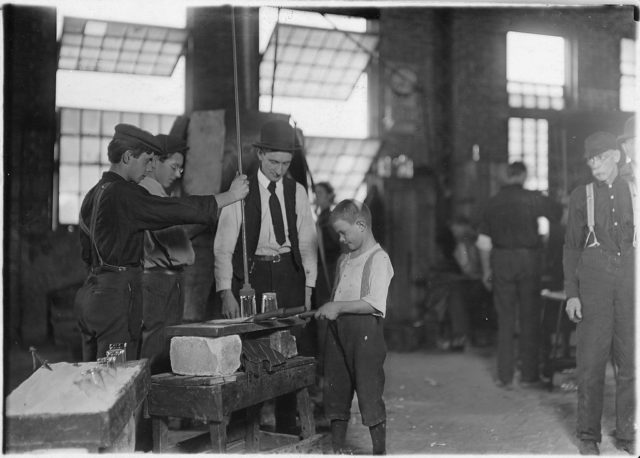 Teaching the young idea. The boss (who began at 10 years of age, and has been at it for 30 years) showing a beginner (who is apparently 9 or 10), October 1908 Phto Credit
