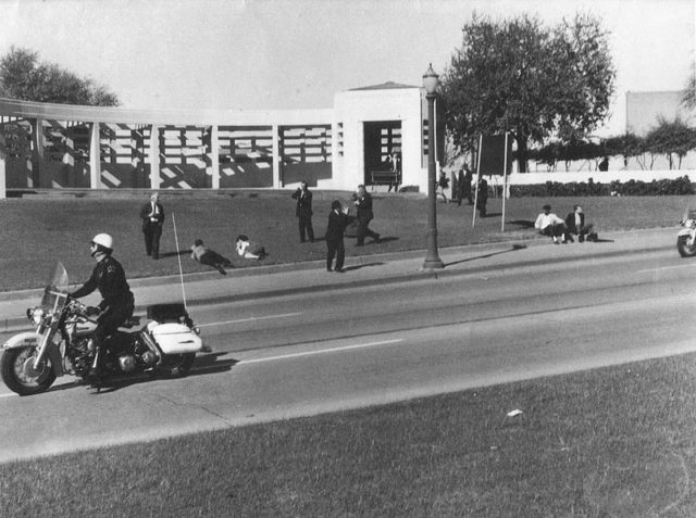 In the aftermath of the assassination, two men can be seen sitting together on the sidewalk on the right side of the photograph. The “umbrella man” is the one in the dark jacket, farthest to the right.