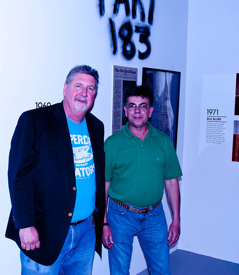 TAKI 183 (right) at a gallery event in 2010, with his tag visible on the wall behind him. Photo Credit