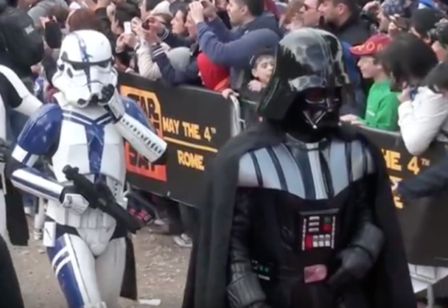 Members of the 501st Legion cosplaying as a stormtrooper (left) and Darth Vader parade in Rome on Star Wars Day 2014. Note the background banner with the May the 4th pun on the phrase May the Force be with you. Photo Credit