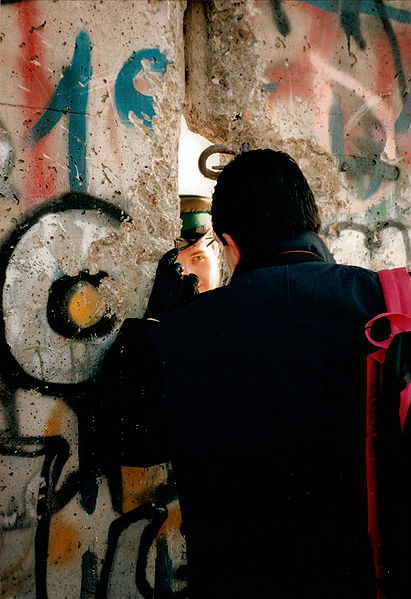 Fall of the Berlin Wall, November 1989. An Eastern guard speaks to a Westerner through a broken seam in the wall. Both were smiling, representing the jubilant spirit of the day. Photo Credit