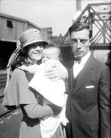 Informal portrait of Buster Keaton and Natalie Talmadge standing with their baby, Joseph Keaton, on the platform of a train station with a bridge in the background in Chicago, Illinois.