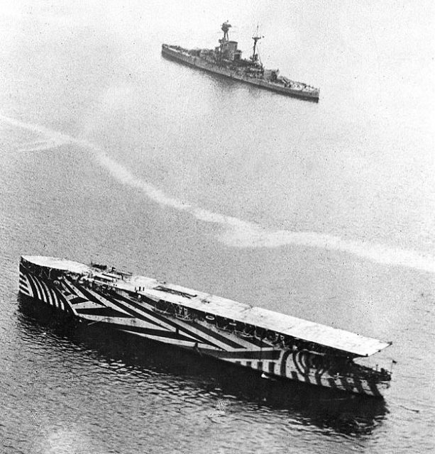 HMS Argus (I49) in a harbor in 1918, painted in dazzle camouflage, with a Renown-class battlecruiser in the distance