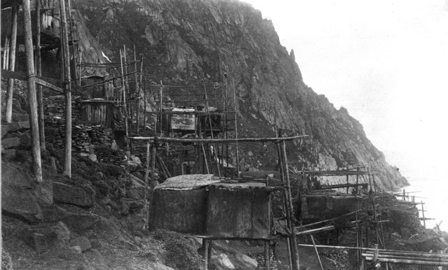 View of Ukivok houses, c 1892. Stilt houses on a rocky bank, built of driftwood poles, planks, and hide or fabric covers. Some stone structures also visible as well as a few residents