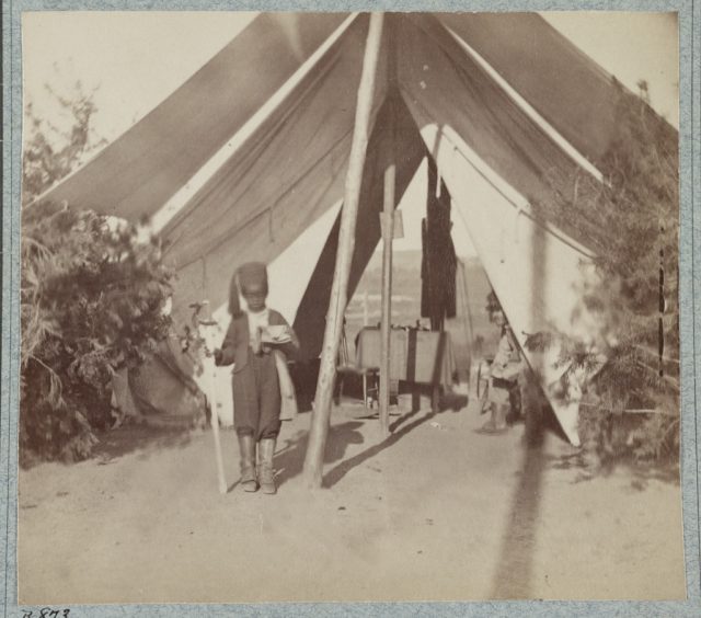 Photograph shows young African American camp servant of Colonel Lloyd Aspinwall of Field & Staff, 22nd New York Infantry Regiment, standing in front of tent with a sword as Aspinwall peeks from inside the tent. Photo Credit