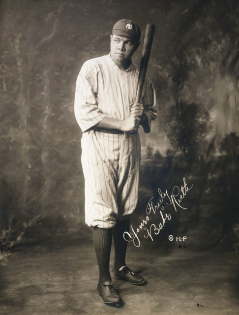 Babe Ruth, full-length portrait, in his baseball outfit holding a bat. His signature on the image reads “Yours truly ‘Babe’ Ruth.”