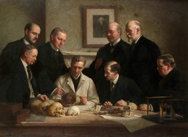 Group portrait of the Piltdown skull being examined. Back row (from left): F. O. Barlow, G. Elliot Smith, Charles Dawson, Arthur Smith Woodward. Front row: A. S. Underwood, Arthur Keith, W. P. Pycraft, and Ray Lankester. Note the portrait of Charles Darwin on the wall. Painting by John Cooke, 1915.