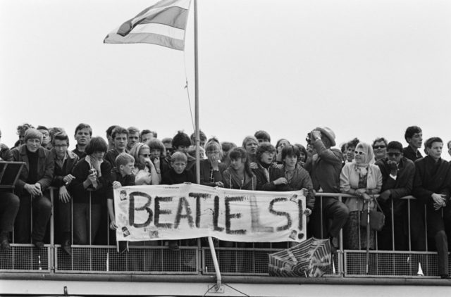 Young fans await their idols at the arrival of The Beatles at Schiphol. The banner makers have probably tried to fix an initial misspelling. Photo Credit