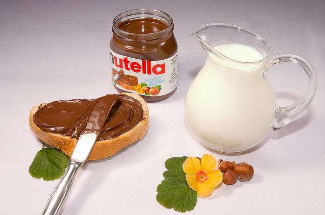 Nutella in its jar and also spread on bread, along with hazelnuts and a pitcher of milk. Author: donald. CC BY-SA 3.0