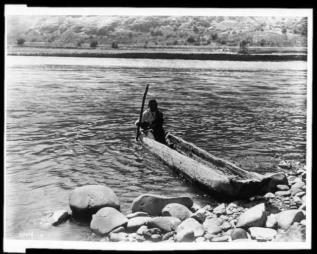 Nez Percé man with pole maneuvering dugout canoe to rocky shore. Author Edward S Curtis. Library of Congress