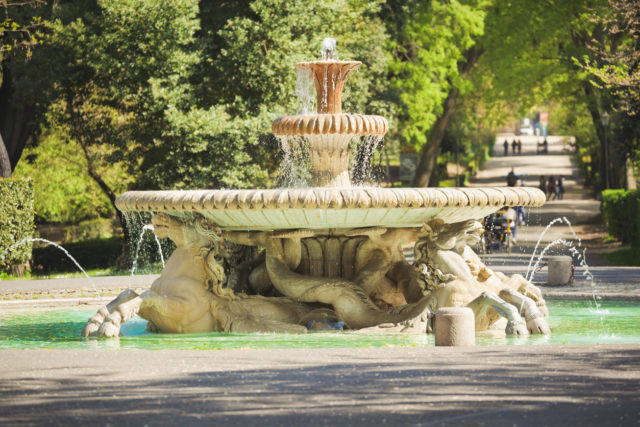 Old (Baroque Style) fountain in the Roman park. In the background park alley with walking people.