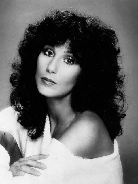 Cher with black curly hair, wearing a white dress