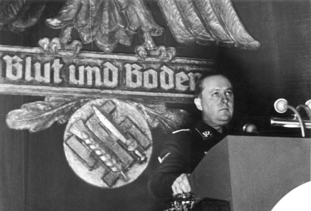Richard Walther Darré addressing a meeting between the farming community in Goslar on 13 December 1937, standing in front of a Reichsadler and Swastika crossed with a sword and a wheat sheaf labeled “Blood and Soil” (from the German Federal Archive). Photo credit Bundesarchiv, Bild CC BY-SA 3.0 de