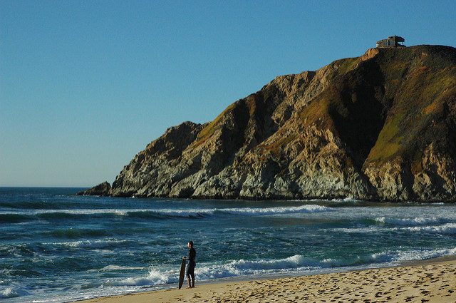 Surfer standing on the beach with Devil’s Slide bunker in the top right – Author: Wonderlane – CC BY 2.0