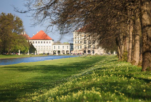 Munich, Germany – April 17, 2011: Park with Alley of trees, canal and back side of the baroque Nymphenburg Palace, Munich (completed in 1675). The huge park (490 acre, 200 ha) behind the palace is a popular venue for a sunday walk with the family. 300000 people are visiting the Palace each year. The canal runs through the whole park and even farther through Munich.