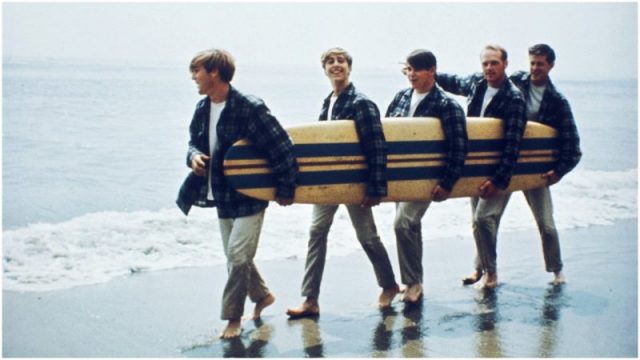 Rock and roll band The Beach Boys walk along the beach holding a surfboard for a portrait session in August 1962 in Los Angeles, California. (L-R) Dennis Wilson, David Marks, Mike Love, Carl Wilson, Brian Wilson. Photo by Michael Ochs Archives/Getty Images