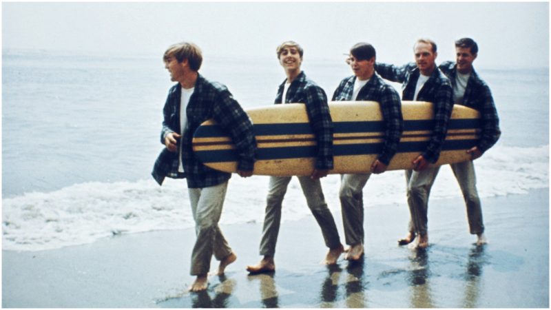 Rock and roll band The Beach Boys walk along the beach holding a surfboard for a portrait session in August 1962 in Los Angeles, California. (L-R) Dennis Wilson, David Marks, Mike Love, Carl Wilson, Brian Wilson. (Photo by Michael Ochs Archives/Getty Images)