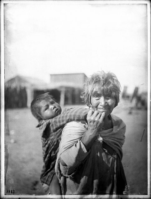 A grandmother, not too old to carry her grandchild on her back. Photo taken at the Palomas Indian Reservation, c. 1900.